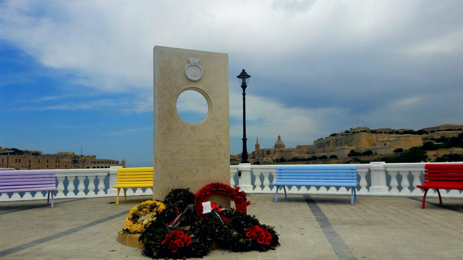 A stone memorial on a seafront promenade in Malta, with coloured benches and the Valletta skyline behind.